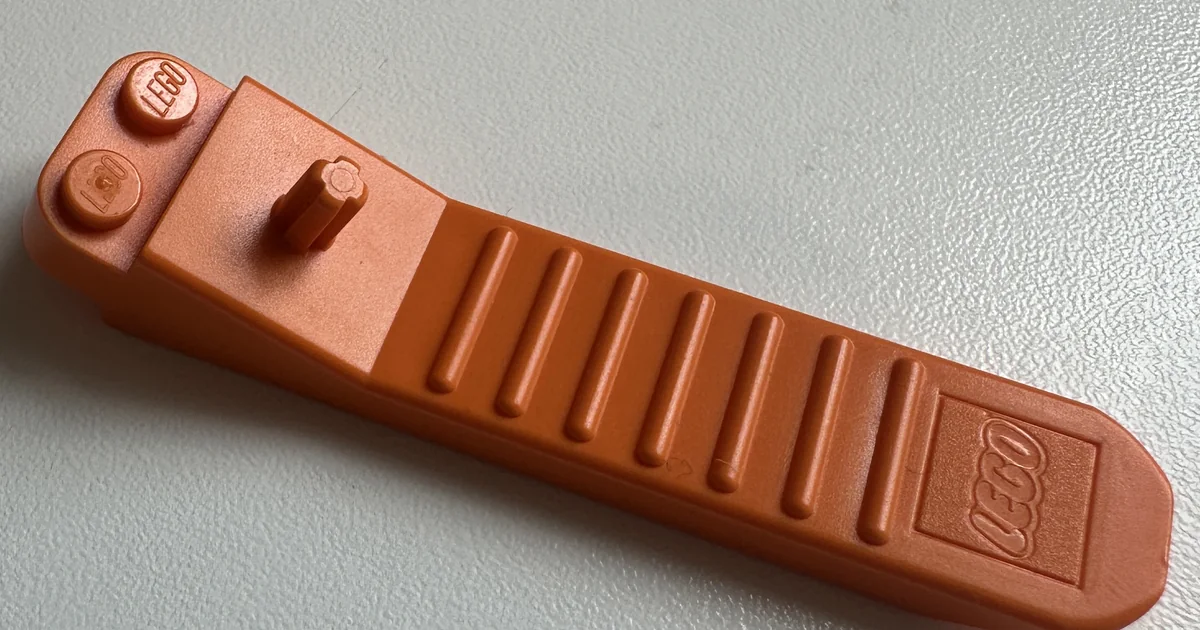 An orange LEGO brick separator lying on a white surface, with two studs and a short upright axle at one end, a ridged grip along the middle, and a thin tapered wedge at the other end.