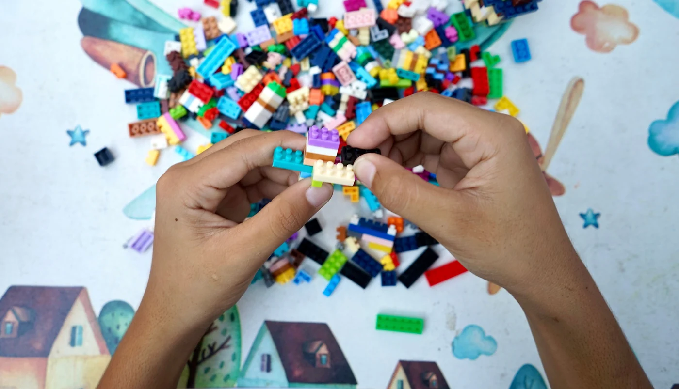 Two hands joining small LEGO bricks together above a scattered pile of mixed pieces on a table.